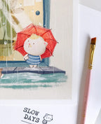 A close-up lifestyle photo of the rainy day cat art print, focusing on the character holding the red umbrella. A pink paintbrush sits to the right on a textured white background.