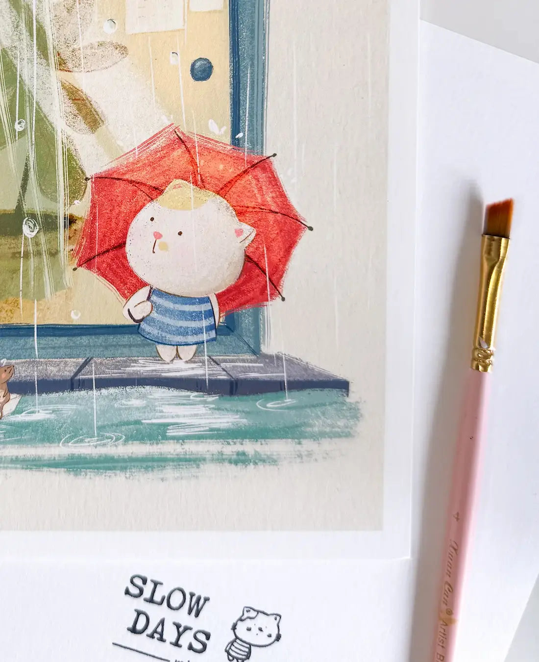 A close-up lifestyle photo of the rainy day cat art print, focusing on the character holding the red umbrella. A pink paintbrush sits to the right on a textured white background.