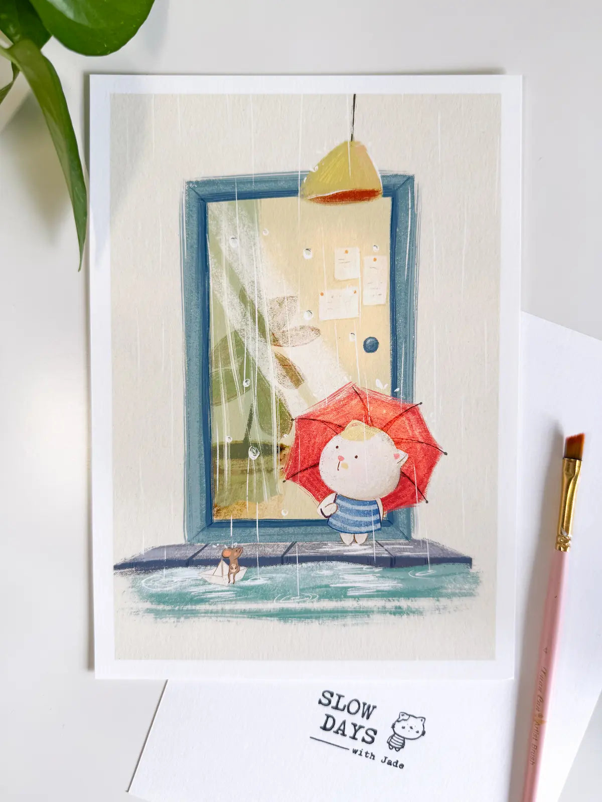 A lifestyle flat lay photo of the rainy day cat art print resting on a white desk with green plant leaves at the top left. A pink artist's paintbrush sits along the right side of the print, showcasing the textured white border and cozy rain scene.