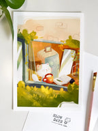 A lifestyle flat lay photo of the window cat in a slow morning theme  art print. The print lies flat on a white surface with a pink artist's paintbrush along the right border and green plant leaves framing the top-left corner.
