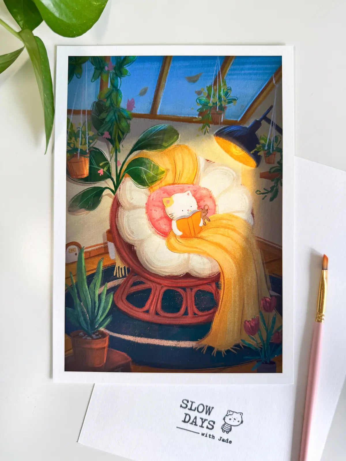 A lifestyle flat lay photo of the greenhouse reading cat art print featuring richer, deeper shadow tones. The printed artwork sits on a white desk backdrop with a pink paintbrush on the right side and a green leaf at the top left.