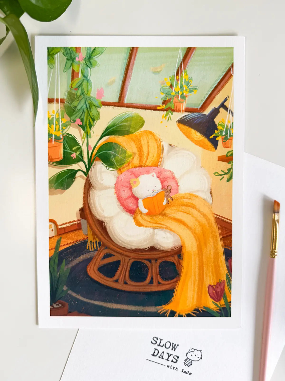 A lifestyle flat lay photo of the greenhouse reading cat art print sitting on a clean white surface. The scene is styled with green houseplant leaves stretching in from the top left and a pink artist's paintbrush on the right.