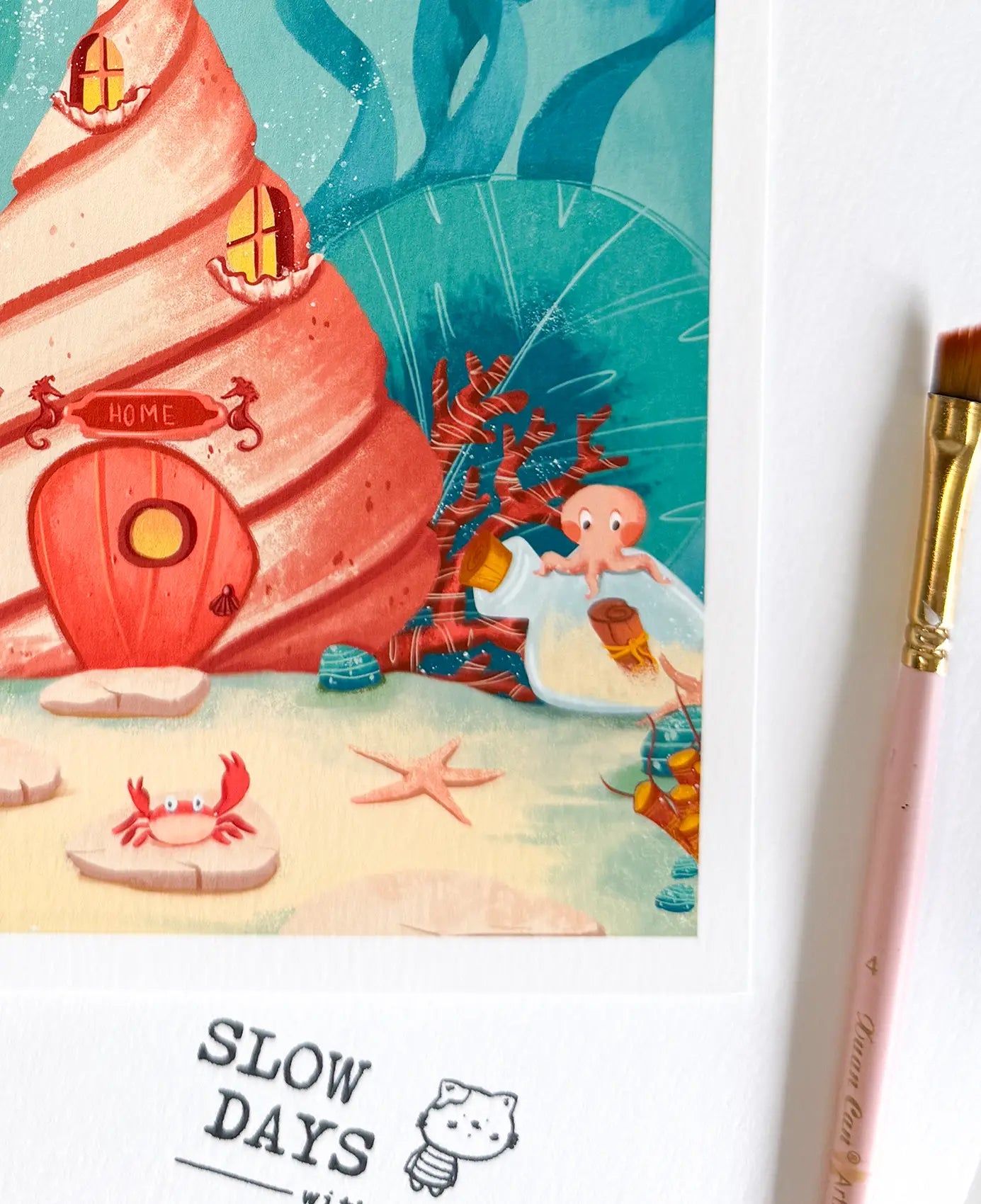 A close-up lifestyle photo focusing on the bottom right corner of the conch shell art print, showcasing a tiny orange octopus sitting on a message in a bottle. A pink artist's paintbrush sits to the right.