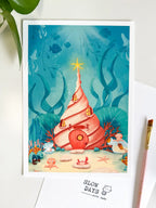 A lifestyle flat lay photo of the conch shell underwater art print styled on a white desk. Green leaves frame the top left corner, while a pink paintbrush and a white "Slow Days" branded card sit along the bottom and right edges.