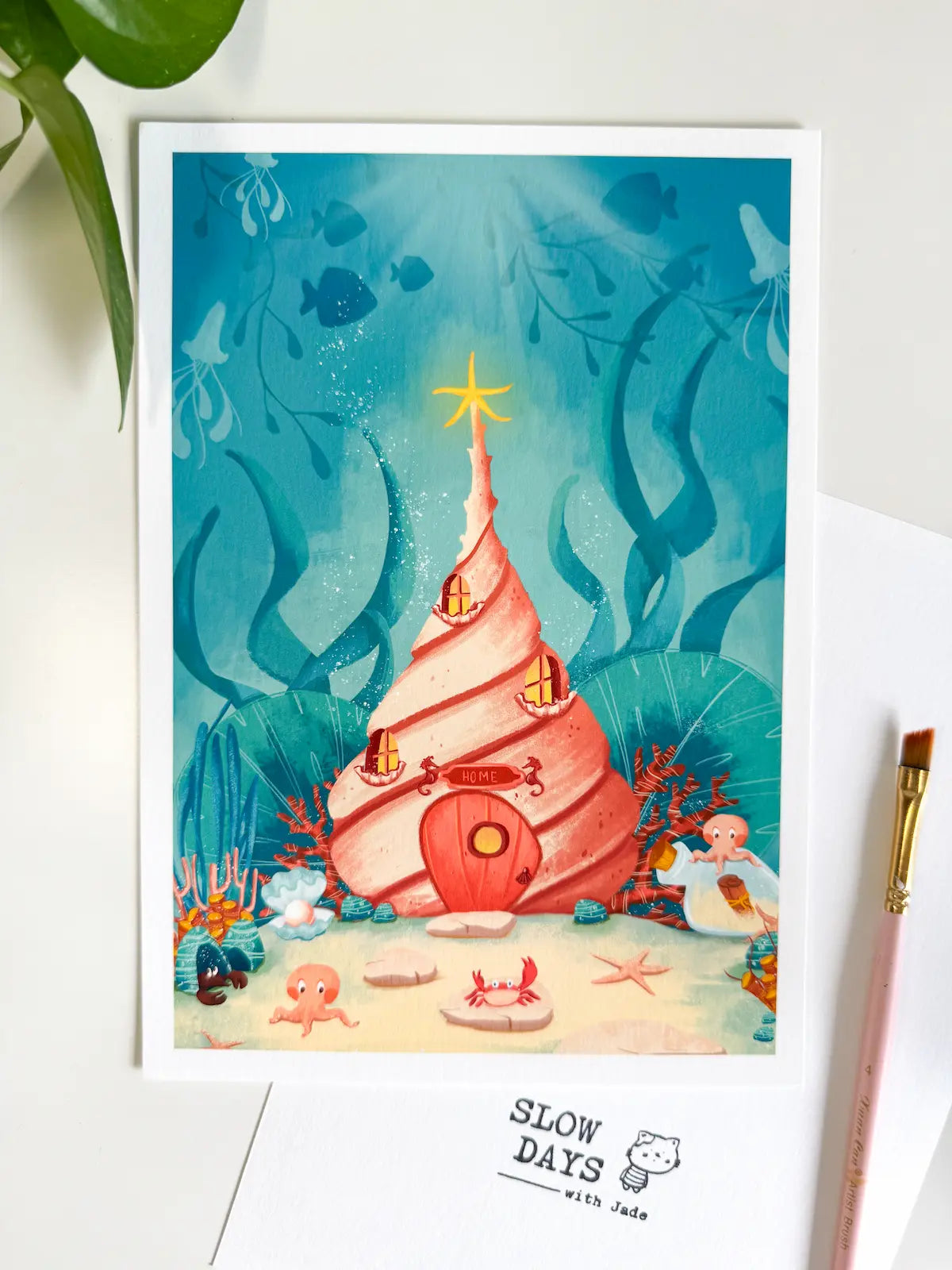 A lifestyle flat lay photo of the conch shell underwater art print styled on a white desk. Green leaves frame the top left corner, while a pink paintbrush and a white "Slow Days" branded card sit along the bottom and right edges.