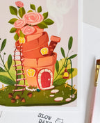 A close-up lifestyle photo of the terracotta flowerpot house art print, showing the textured paper details. A pink artist's paintbrush rests on the right side of the frame next to the print.