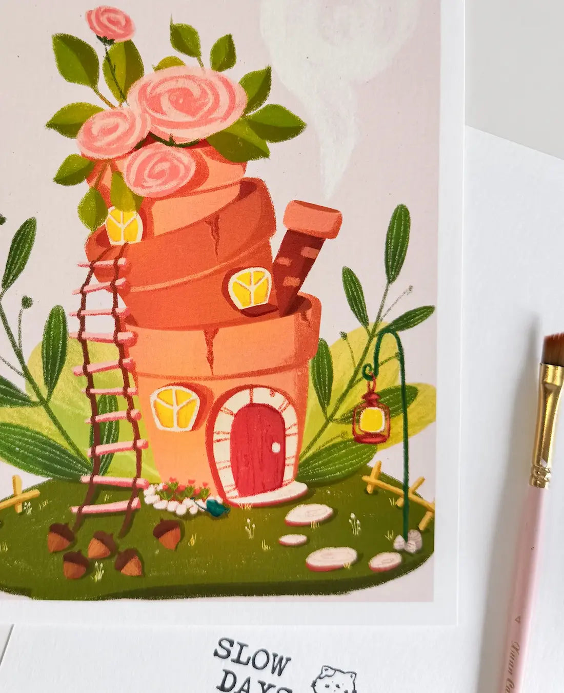 A close-up lifestyle photo of the terracotta flowerpot house art print, showing the textured paper details. A pink artist's paintbrush rests on the right side of the frame next to the print.