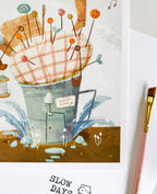 A close-up lifestyle photo of the pincushion house art print, highlighting the fine texture of the plaid cushion and colorful pins. A pink artist's paintbrush rests vertically along the right border.