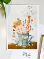 A lifestyle flat lay photo of the pincushion house art print resting on a clean white desk setup. Styled with a fresh green plant in the upper left corner, a pink artist's paintbrush on the right, and a "Slow Days" logo card below.