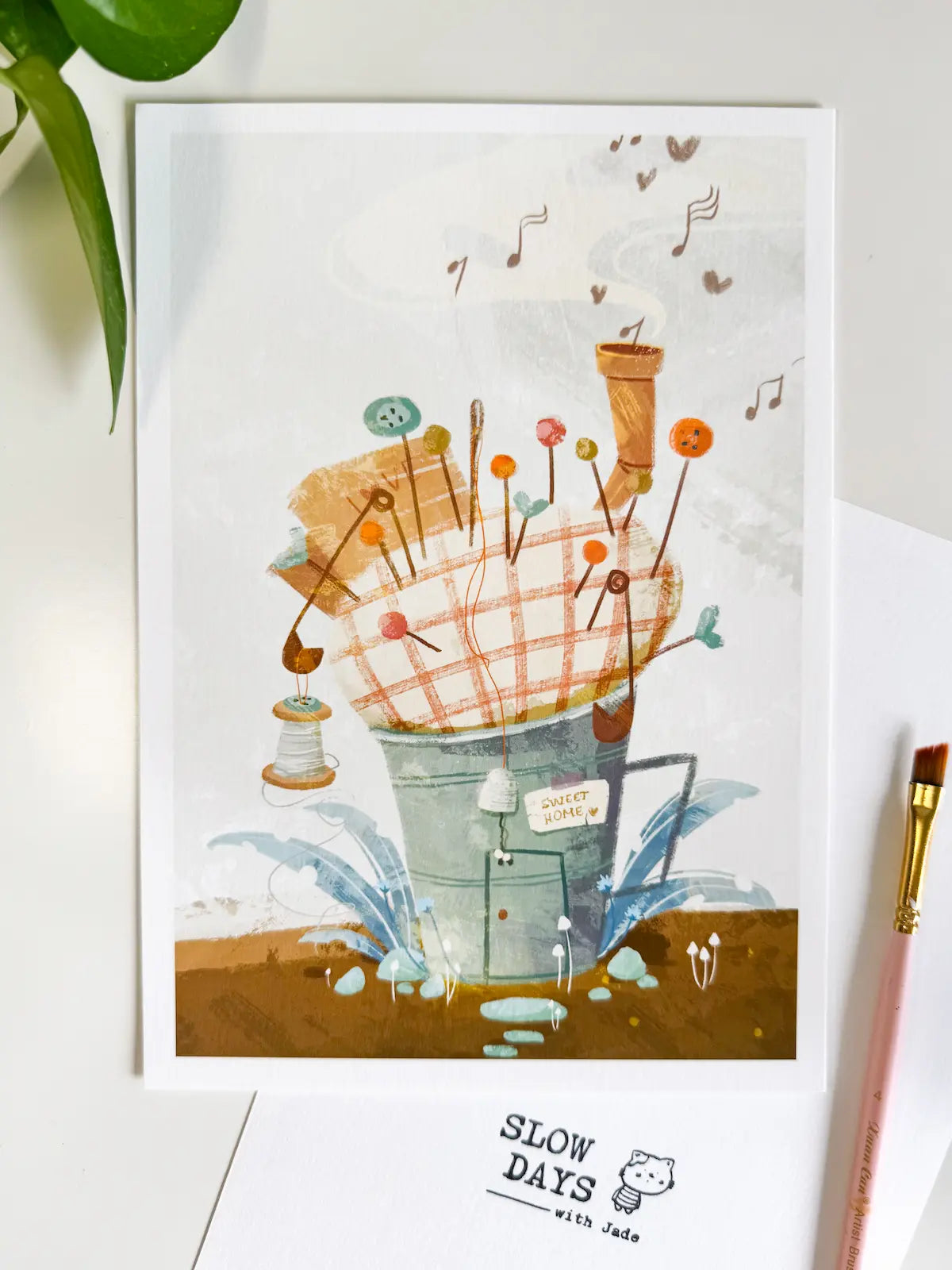A lifestyle flat lay photo of the pincushion house art print resting on a clean white desk setup. Styled with a fresh green plant in the upper left corner, a pink artist's paintbrush on the right, and a "Slow Days" logo card below.