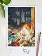 A lifestyle flat lay photo of the forest fairy art print laid out on a white surface. The print is framed by green plant leaves at the top left, with a pink paintbrush along the right side and a "Slow Days" card at the bottom.
