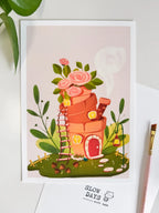 A lifestyle flat lay photo of the flowerpot house art print resting on a white table. The scene includes green plant leaves peaking in from the top left and a pink paintbrush on the right side.