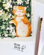 A close-up lifestyle photo of the orange tabby cat art print, showcasing the textured paper and fine lines. A pink artist's paint brush lies along the right edge of the white border.