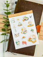 A full lifestyle flat lay photo of the "Slow Morning" sticker sheet resting on a dark brown textured leather journal with an orange leather closure. A green leafy plant stem frames the left side against a white background, displaying the entire sheet of morning-themed cat stickers.