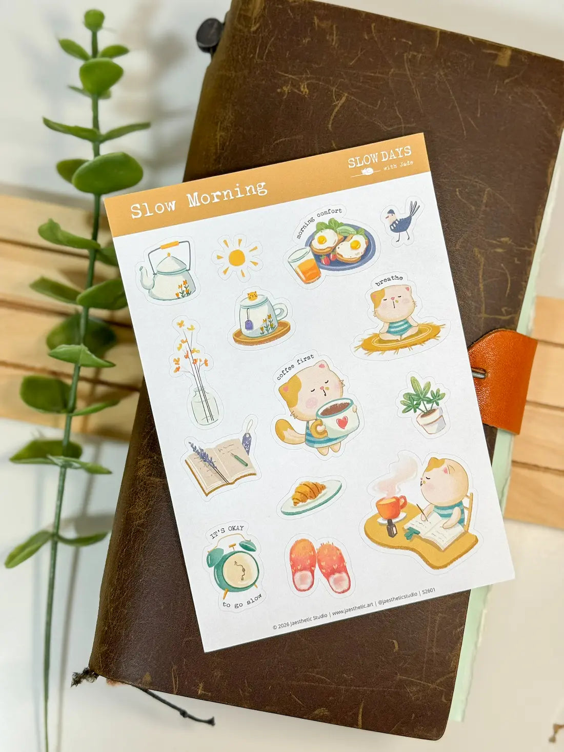 A full lifestyle flat lay photo of the "Slow Morning" sticker sheet resting on a dark brown textured leather journal with an orange leather closure. A green leafy plant stem frames the left side against a white background, displaying the entire sheet of morning-themed cat stickers.