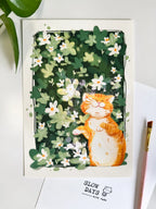 A lifestyle flat lay photo of the happy orange tabby cat art print on a clean white desk, framed by green houseplant leaves at the top left and a pink artist's paint brush on the right.