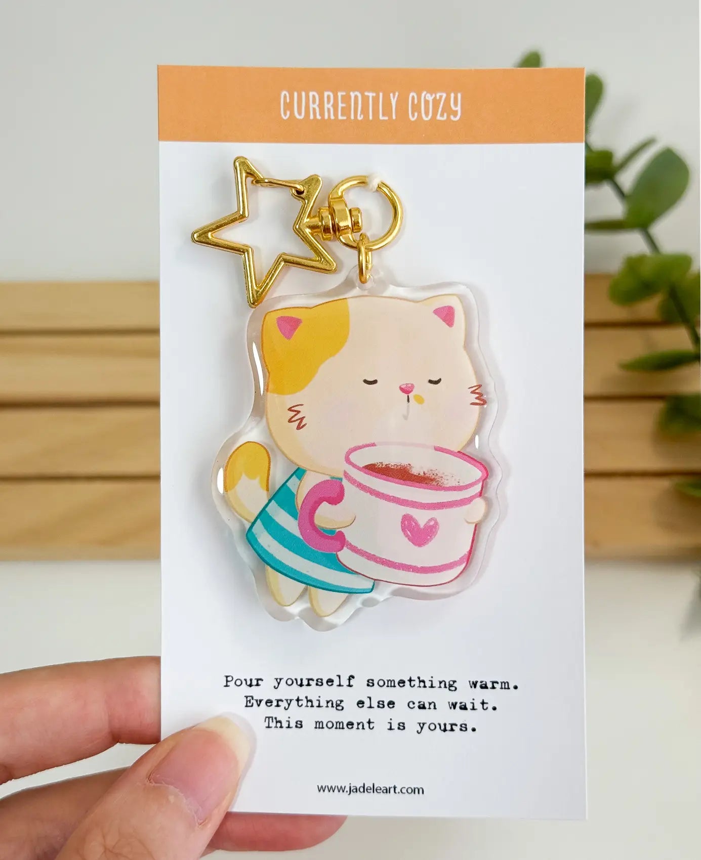 An acrylic keychain featuring a whimsical illustration of a calico cat wearing a blue striped outfit and holding a large white mug with a pink heart, attached to a gold star-shaped clasp. The backing card reads "CURRENTLY COZY" at the top, and "Pour yourself something warm. Everything else can wait. This moment is yours." at the bottom.