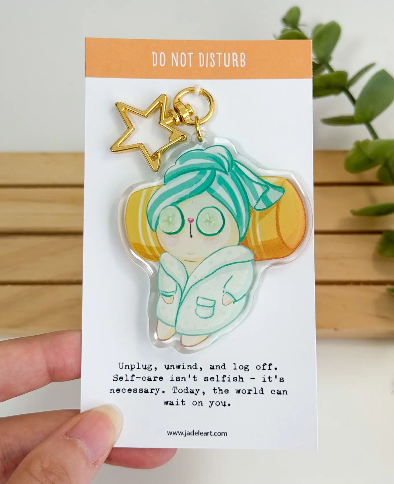 An acrylic keychain featuring a whimsical illustration of a cat wearing a cozy bathrobe, a striped hair towel wrap, and cucumber slices over its eyes while resting against a yellow pillow, attached to a gold star-shaped clasp. The backing card reads "DO NOT DISTURB" at the top, and "Unplug, unwind, and log off. Self-care isn't selfish – it's necessary. Today, the world can wait on you." at the bottom.