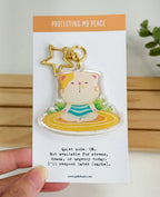 An acrylic keychain featuring a whimsical illustration of a tabby cat meditating on a yellow mat, attached to a gold star-shaped clasp. The backing card reads "Protecting My Peace" at the top, and "Quiet mode: ON. Not available for stress, drama, or urgency today. I'll respond later (maybe)." at the bottom.