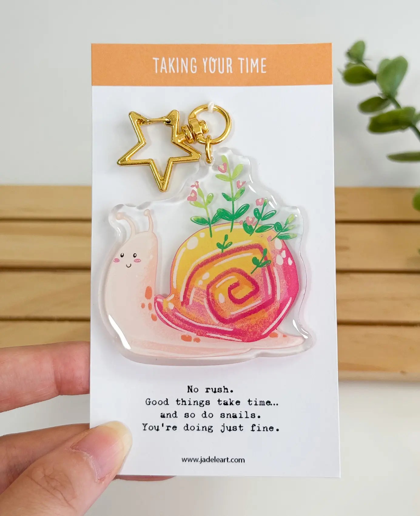 An acrylic keychain featuring a whimsical illustration of a smiling pink snail with green plants growing out of its vibrant pink and yellow shell, attached to a gold star-shaped clasp. The backing card reads "TAKING YOUR TIME" at the top, and "No rush. Good things take time... and so do snails. You're doing just fine." at the bottom.