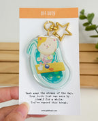 An acrylic keychain featuring a whimsical illustration of a calico cat relaxing in a turquoise bathtub, attached to a gold star-shaped clasp. The backing card reads "OFF DUTY" at the top, and "Wash away the stress of the day. Your to-do list can swim by itself for a while. You've earned this break." at the bottom.