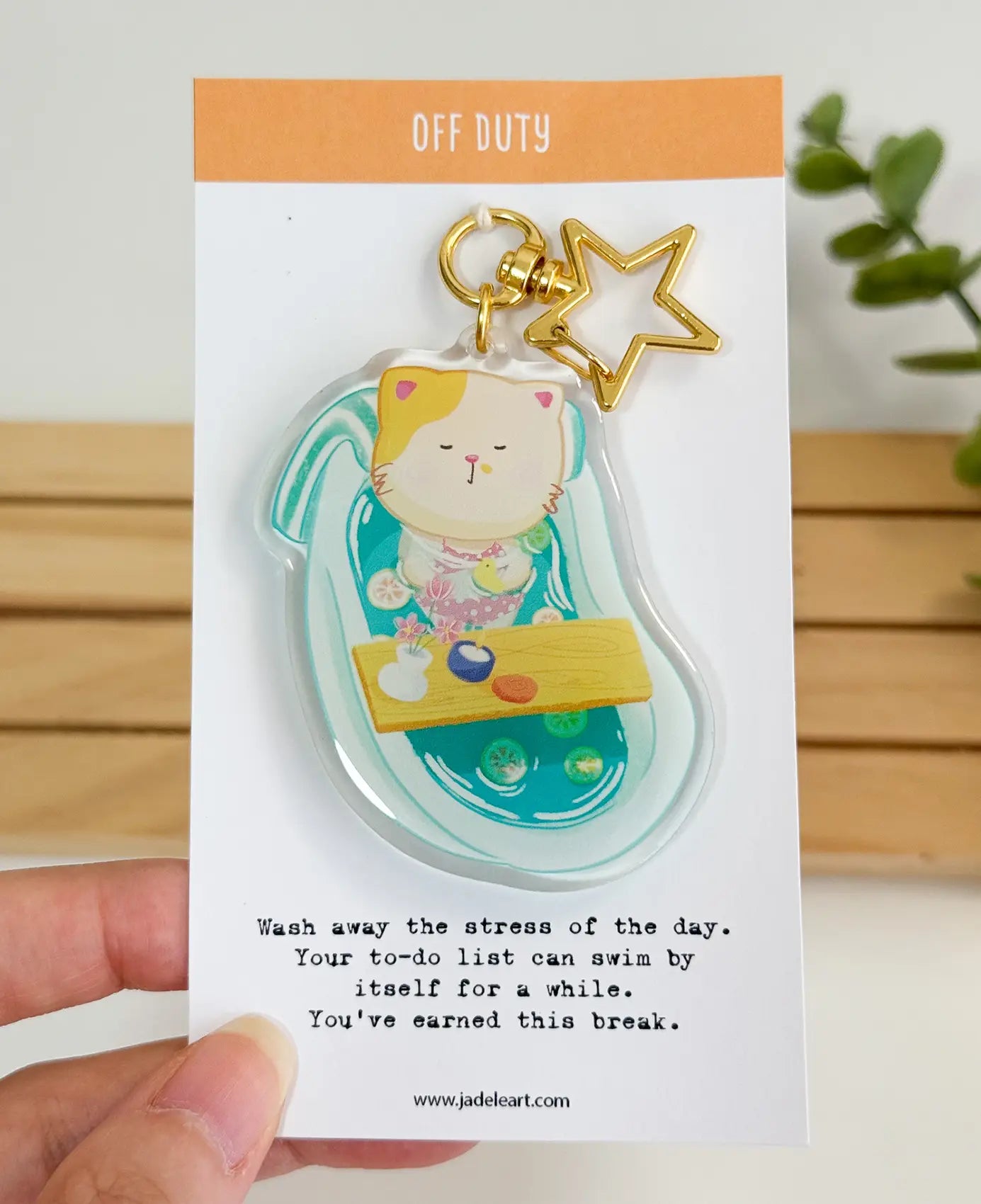 An acrylic keychain featuring a whimsical illustration of a calico cat relaxing in a turquoise bathtub, attached to a gold star-shaped clasp. The backing card reads "OFF DUTY" at the top, and "Wash away the stress of the day. Your to-do list can swim by itself for a while. You've earned this break." at the bottom.