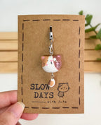A handmade tricolor calico cat phone charm with brown and orange patches and a dangling patterned tail attached to a silver lobster clasp. The clay charm is displayed on a brown kraft paper backing card with a stamped dashed border, a small cat illustration, and the brand name "SLOW DAYS with Jade".