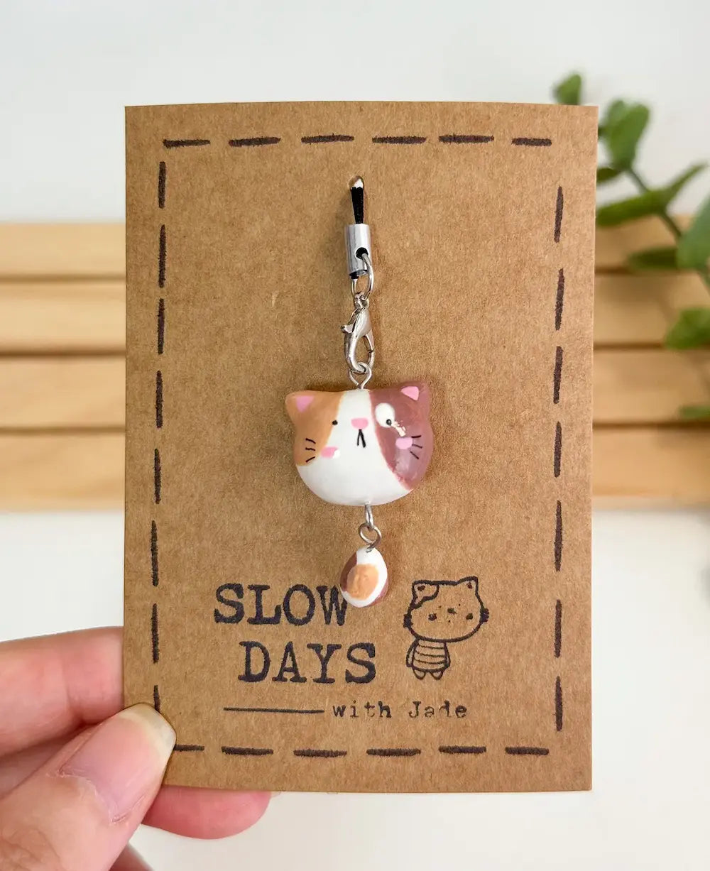 A handmade tricolor calico cat phone charm with brown and orange patches and a dangling patterned tail attached to a silver lobster clasp. The clay charm is displayed on a brown kraft paper backing card with a stamped dashed border, a small cat illustration, and the brand name "SLOW DAYS with Jade".