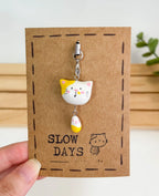 A handmade tabby cat phone charm with a yellow patch over one ear and a dangling yellow and white tail attached to a silver lobster clasp. The clay charm is displayed on a brown kraft paper backing card with a stamped dashed border, a small cat illustration, and the brand name "SLOW DAYS".
