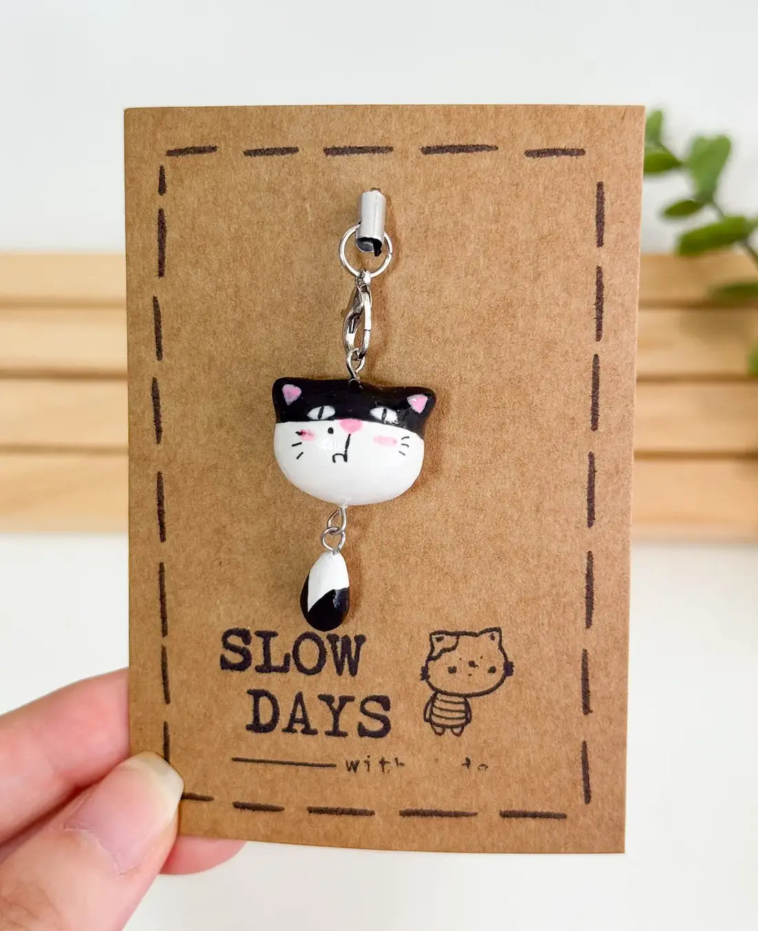 A handmade clay tuxedo cat phone charm with a dangling black-and-white tail attached to a silver lobster clasp. The charm is displayed on a brown kraft paper backing card with a stamped dashed border, a small cat illustration, and the brand name "SLOW DAYS".