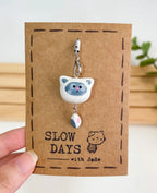A handmade Siamese or point-colored cat phone charm with a dangling gray and pink tail attached to a silver lobster clasp. The clay charm is displayed on a brown kraft paper backing card with a stamped dashed border, a small cat illustration, and the brand name "SLOW DAYS with Jade".