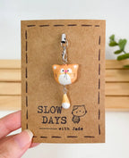 A handmade orange tabby cat phone charm with a dangling orange and white tail attached to a silver lobster clasp. The clay charm is displayed on a brown kraft paper backing card with a stamped dashed border, a small cat illustration, and the brand name "SLOW DAYS with Jade".
