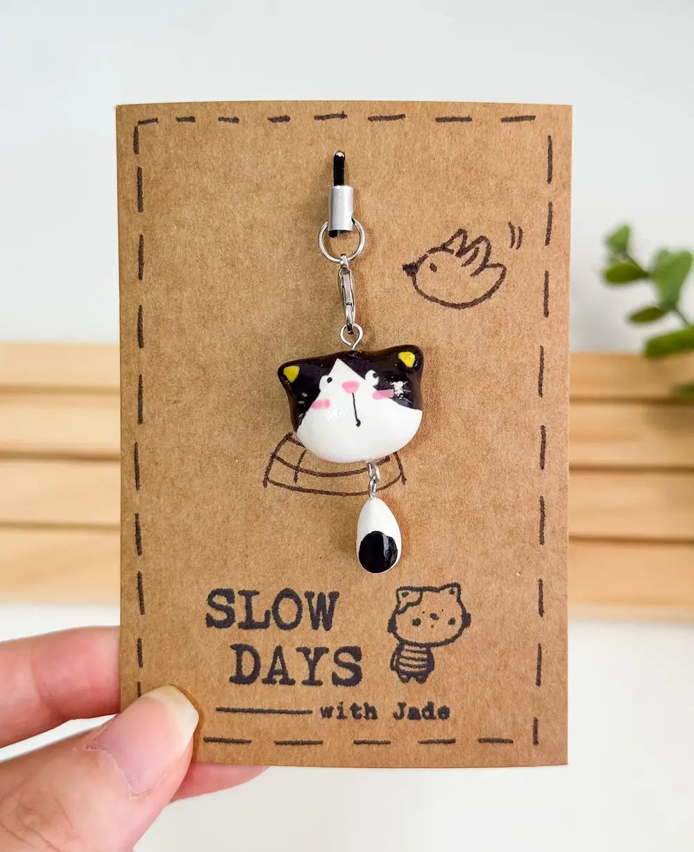 A handmade cow cat or piebald cat phone charm with yellow inner ears and a dangling black and white tail attached to a silver lobster clasp. The clay charm is displayed on a brown kraft paper backing card with a stamped dashed border, a small cat illustration, a little bird sketch, and the brand name "SLOW DAYS with Jade".