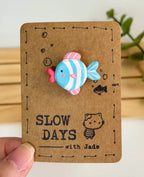 A handmade clay pin shaped like a cute striped fish with a light blue and white body, a bright blue tail, pink dorsal and ventral fins, and bright pink lips. The pin is attached to a brown kraft paper backing card decorated with small stamped bubbles, a tiny fish sketch, a little cat illustration, and the brand name "SLOW DAYS with Jade".