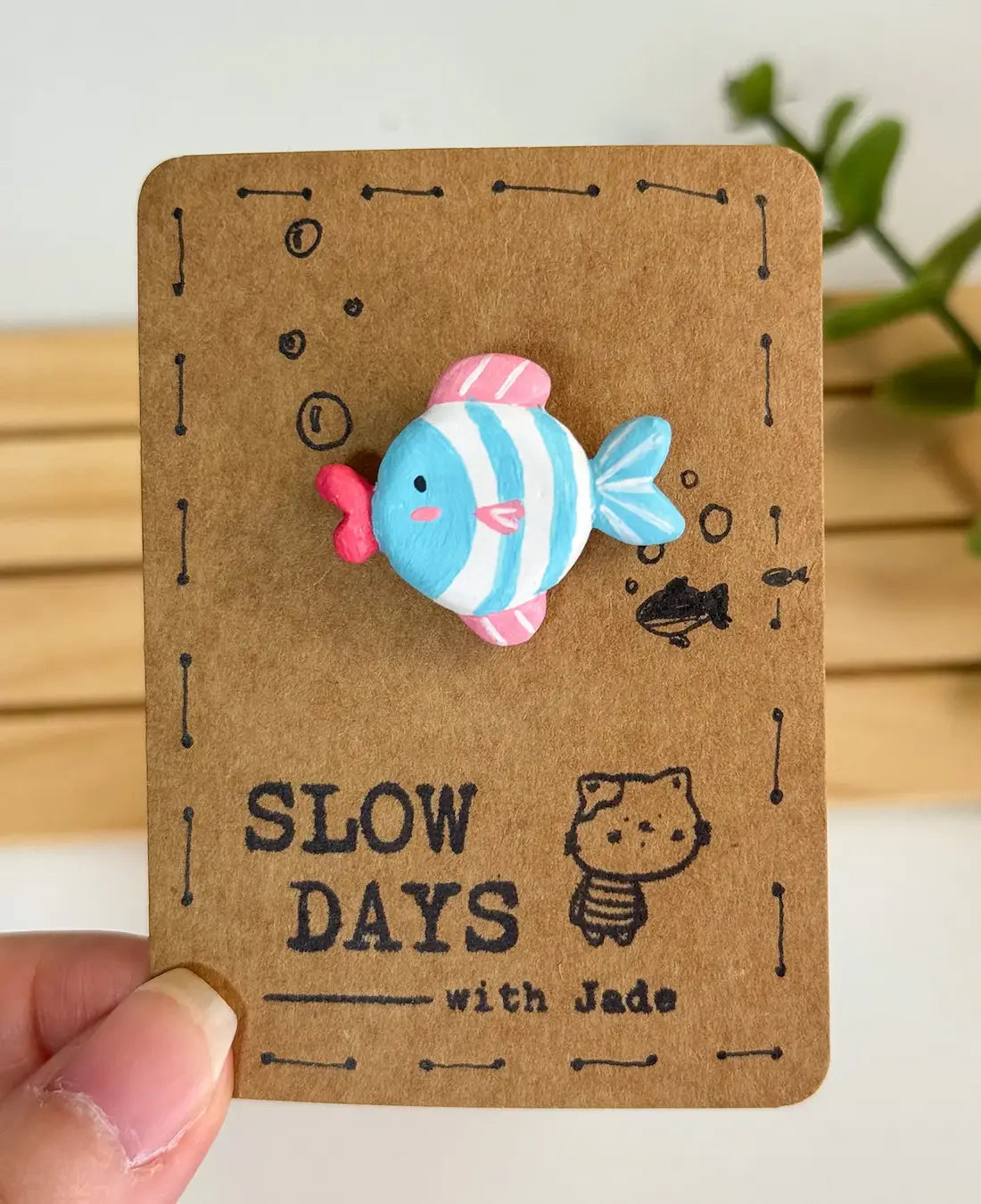 A handmade clay pin shaped like a cute striped fish with a light blue and white body, a bright blue tail, pink dorsal and ventral fins, and bright pink lips. The pin is attached to a brown kraft paper backing card decorated with small stamped bubbles, a tiny fish sketch, a little cat illustration, and the brand name "SLOW DAYS with Jade".