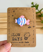 A handmade clay pin shaped like a cute striped fish with a blue and white body, pink fins, and light pink lips. The pin is attached to a brown kraft paper backing card decorated with small stamped bubbles, a tiny fish sketch, a little cat illustration, and the brand name "SLOW DAYS with Jade".