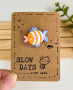 Handmade clay pin of Finn the Fish in soft orange with pastel blue fins, displayed on a brown kraft paper backing card that says Slow Days with Jade.
