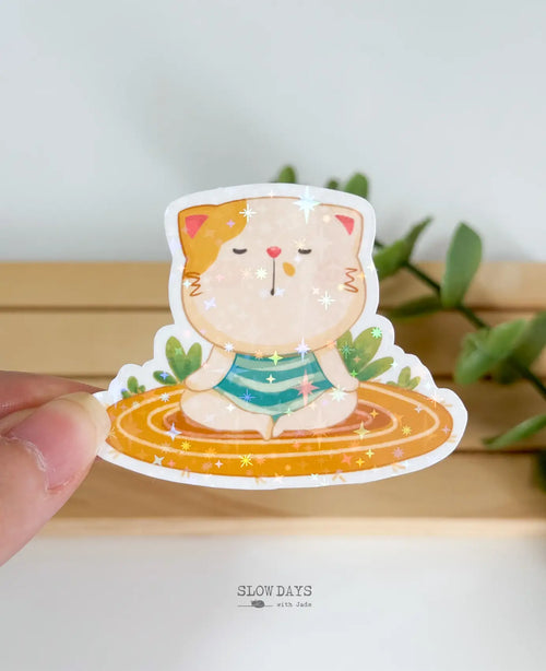 A die-cut waterproof vinyl sticker of a peaceful cream and orange cat meditating with closed eyes on an orange striped rug. The sticker features a bright, multi-colored star holographic sparkle finish