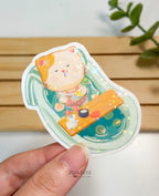 A die-cut waterproof vinyl sticker of a cute cat relaxing in a cozy mint green clawfoot bathtub filled with water, citrus slices, and pink flowers. A little yellow rubber duck sits on a wooden bath tray, covered in a star holographic finish.