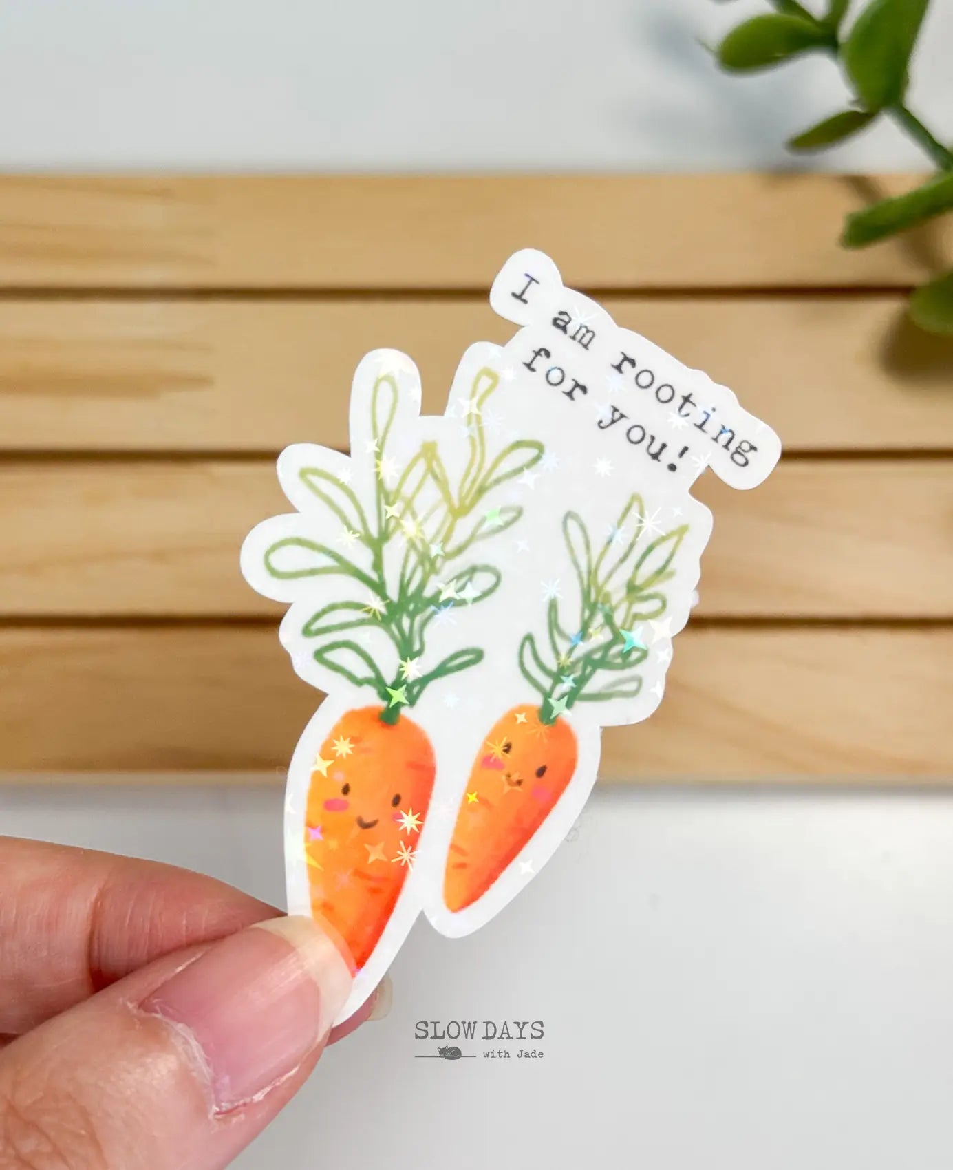 A die-cut waterproof vinyl sticker featuring two illustrated smiling carrots with green leafy tops. Above them, typewriter-style text reads, "I am rooting for you!" The sticker is overlaid with a star holographic sparkle effect.