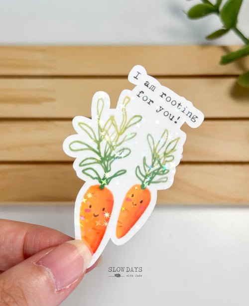 A die-cut waterproof vinyl sticker featuring two illustrated smiling carrots with green leafy tops. Above them, typewriter-style text reads, "I am rooting for you!" The sticker is overlaid with a star holographic sparkle effect.