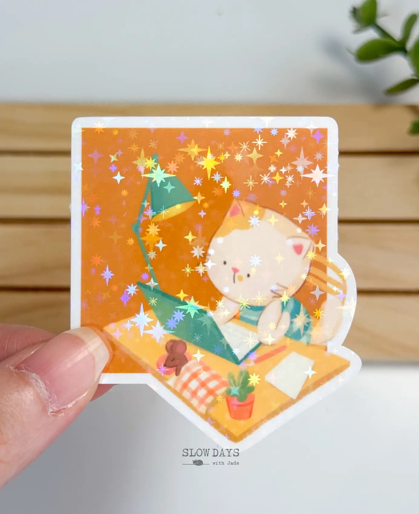 A hand-held close-up of a die-cut waterproof vinyl sticker showing a cute cream and orange cat working diligently at a desk. The cozy workspace illustration includes an open teal laptop, an adjustable desk lamp, a small potted plant, and a tiny sleeping mouse tucked into a checkered blanket, all coated in a brilliant star holographic sparkle finish.