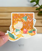 A die-cut waterproof vinyl sticker showing a cream and orange cat writing in a notebook at a yellow desk. The cozy scene includes an open laptop, a red mug, a teal desk lamp, and a small potted plant, coated in a vivid star holographic sparkle.