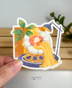 A die-cut waterproof vinyl sticker featuring a cozy scene of a small cat tucked into a round papasan chair, reading an open book under a long yellow blanket. The background includes a large green potted plant and a dark blue floor lamp, all finished with a star holographic sparkle overlay.