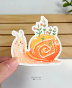A hand-held close-up of a die-cut waterproof vinyl sticker featuring a charming, smiling pink snail. Green leafy stems and small pink flowers sprout from its vibrant orange, swirled shell, completely finished with a multi-colored star holographic sparkle effect.