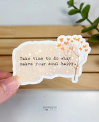 A die-cut waterproof vinyl sticker with a textured beige background featuring a delicate brown branch with small orange heart-shaped leaves. Typewriter text reads, "Take time to do what makes your soul happy," finished with a star holographic sheen.