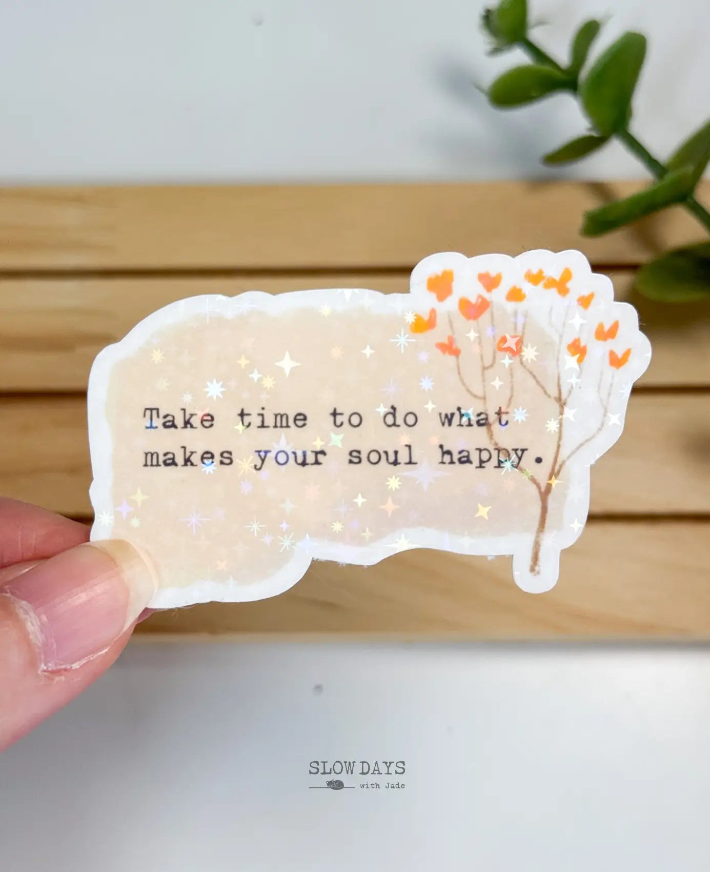 A die-cut waterproof vinyl sticker with a textured beige background featuring a delicate brown branch with small orange heart-shaped leaves. Typewriter text reads, "Take time to do what makes your soul happy," finished with a star holographic sheen.