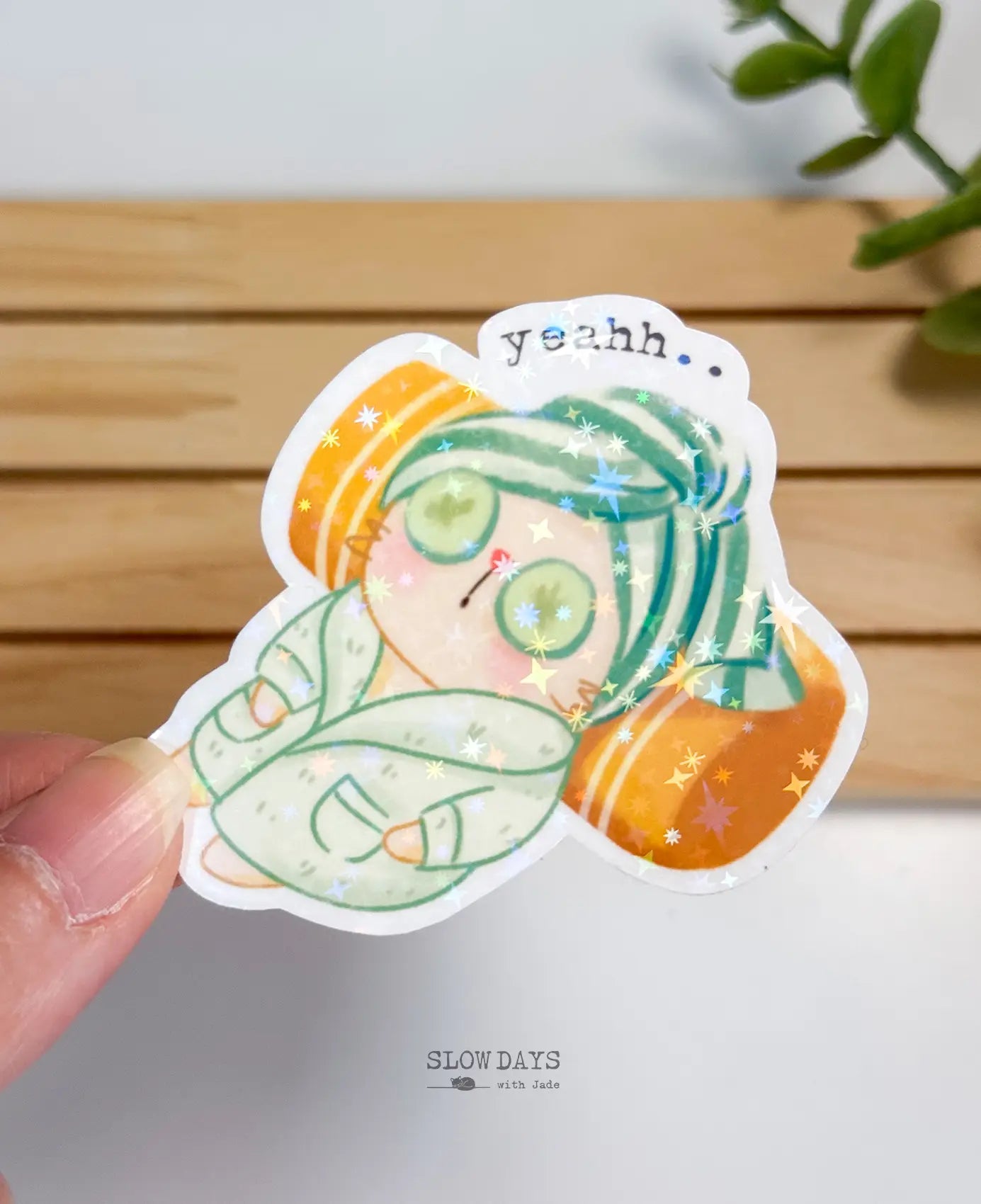 A die-cut waterproof vinyl sticker of a relaxed cat in a mint green bathrobe and matching hair towel wrap with cucumber slices over its eyes, resting on an orange pillow. Small text above reads "yeahh.." with a star holographic sparkle finish.
