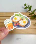 A die-cut waterproof vinyl sticker of a breakfast plate featuring two sunny-side-up eggs on toast, sliced avocado, lettuce, and small red tomatoes, paired with a glass of orange juice. The sticker has a star holographic sparkle overlay.