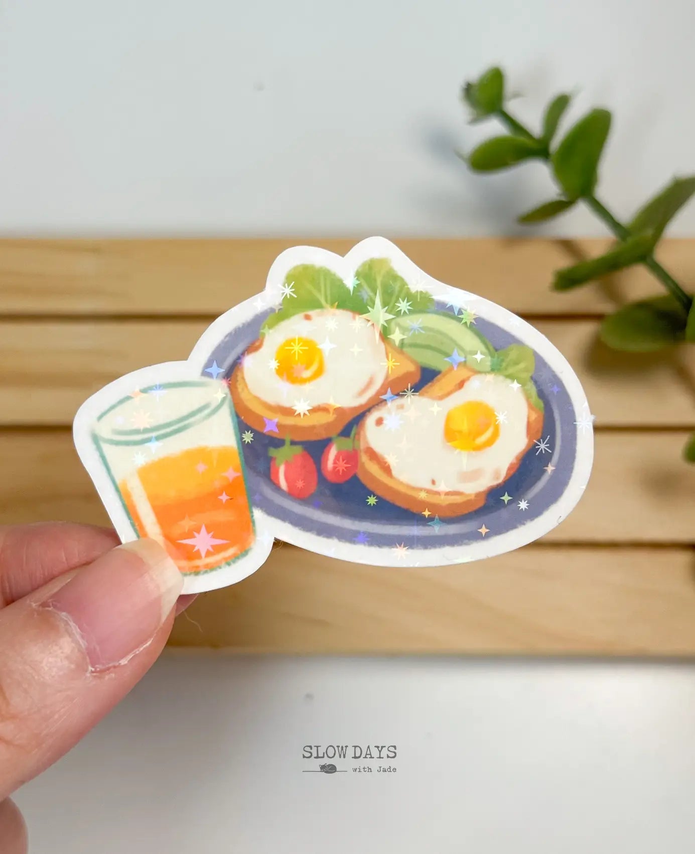 A die-cut waterproof vinyl sticker of a breakfast plate featuring two sunny-side-up eggs on toast, sliced avocado, lettuce, and small red tomatoes, paired with a glass of orange juice. The sticker has a star holographic sparkle overlay.